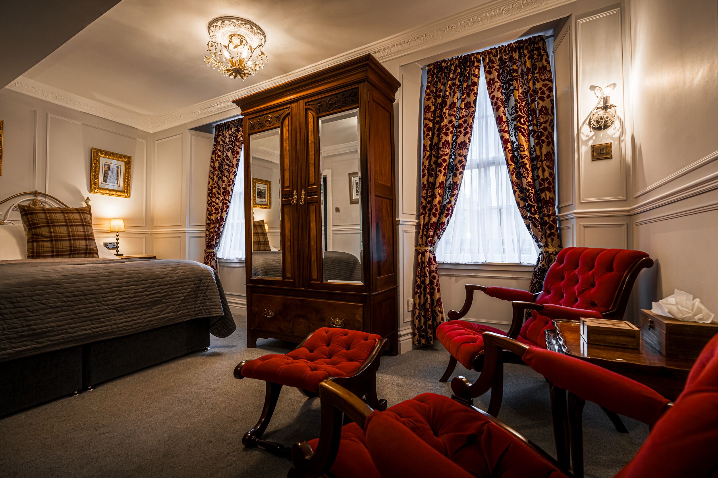 Quarto de hotel com uma cama grande, um guarda-roupa de madeira, uma poltrona vermelha e um banquinho vermelho.