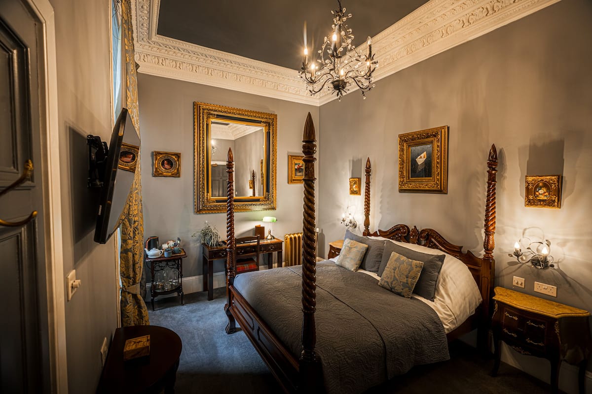 Une chambre à coucher avec un lit à baldaquin, un lustre, des miroirs décoratifs et des œuvres d’art encadrées accrochées aux murs.