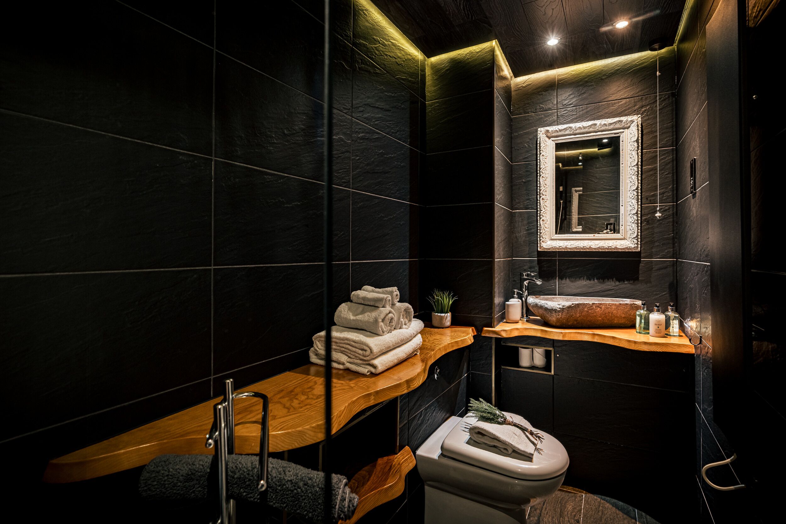 A modern bathroom with dark tiles, a wooden vanity, a sink with a mirror, and a toilet.