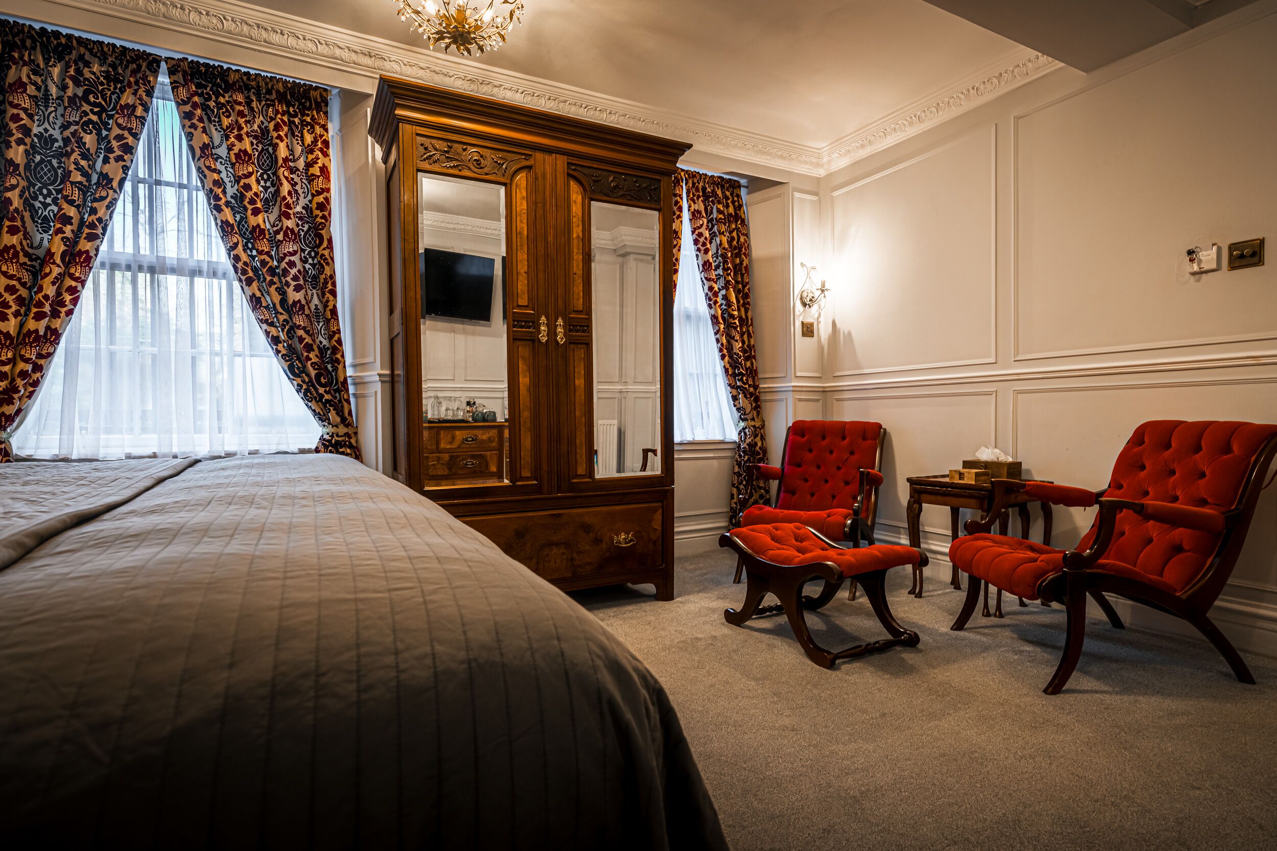 A hotel room with a large bed, a wooden wardrobe, two red armchairs, and a small table.