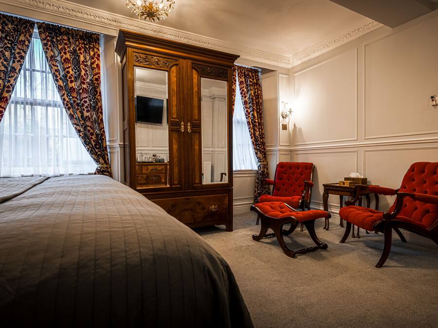 Quarto de hotel com uma cama grande, um guarda-roupa de madeira, duas poltronas vermelhas e uma mesinha.