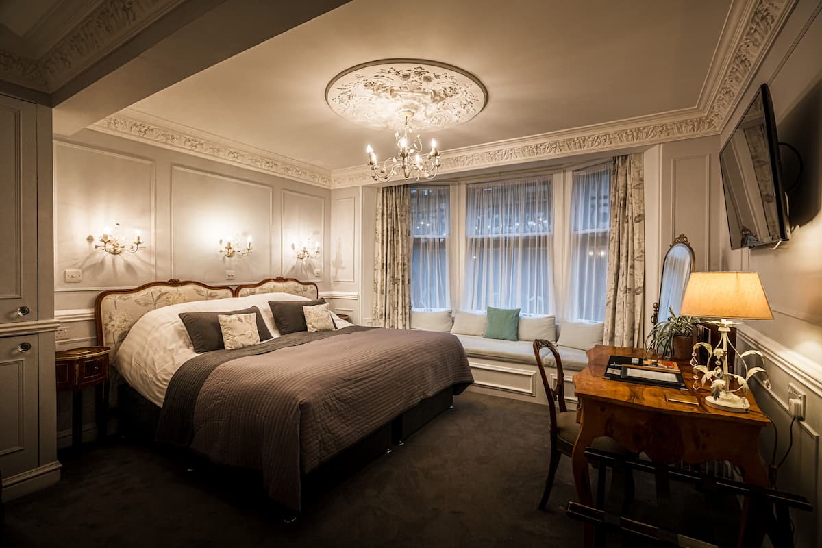 Une chambre élégamment aménagée, dotée d’un grand lit, d’un lustre, d’un bureau avec une lampe et d’une fenêtre avec des rideaux.