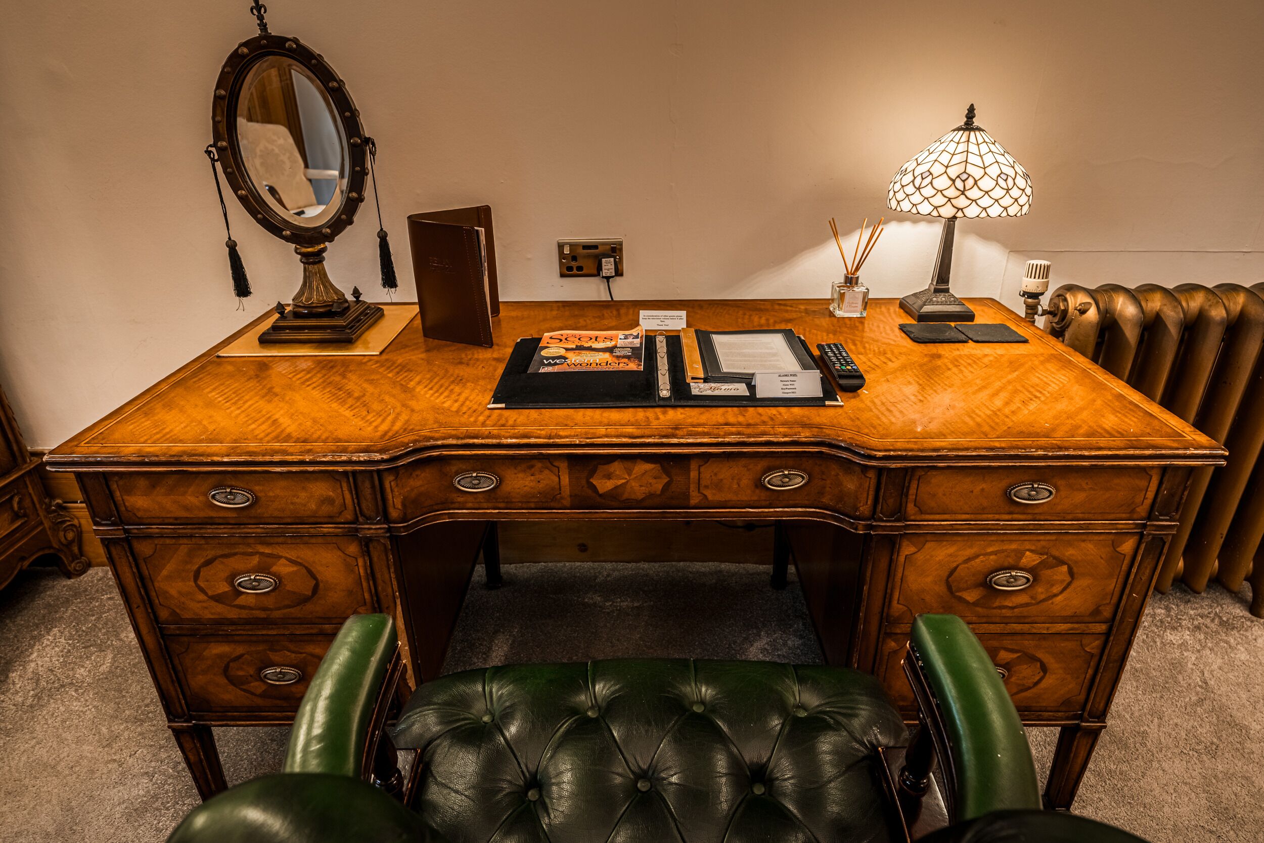 A wooden desk with drawers, a mirror, a lamp, and a green leather chair.