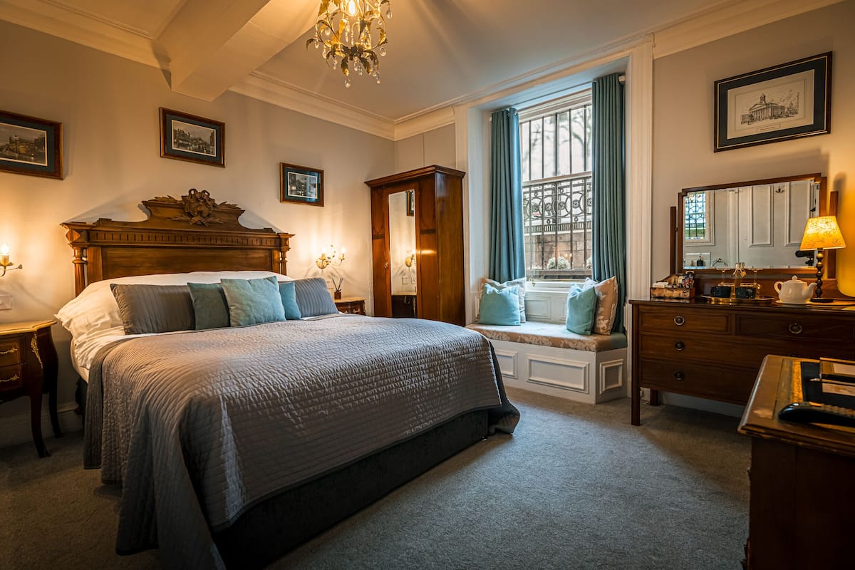 Une chambre à coucher avec un grand lit, une commode, une coiffeuse avec un miroir et une fenêtre avec des rideaux.