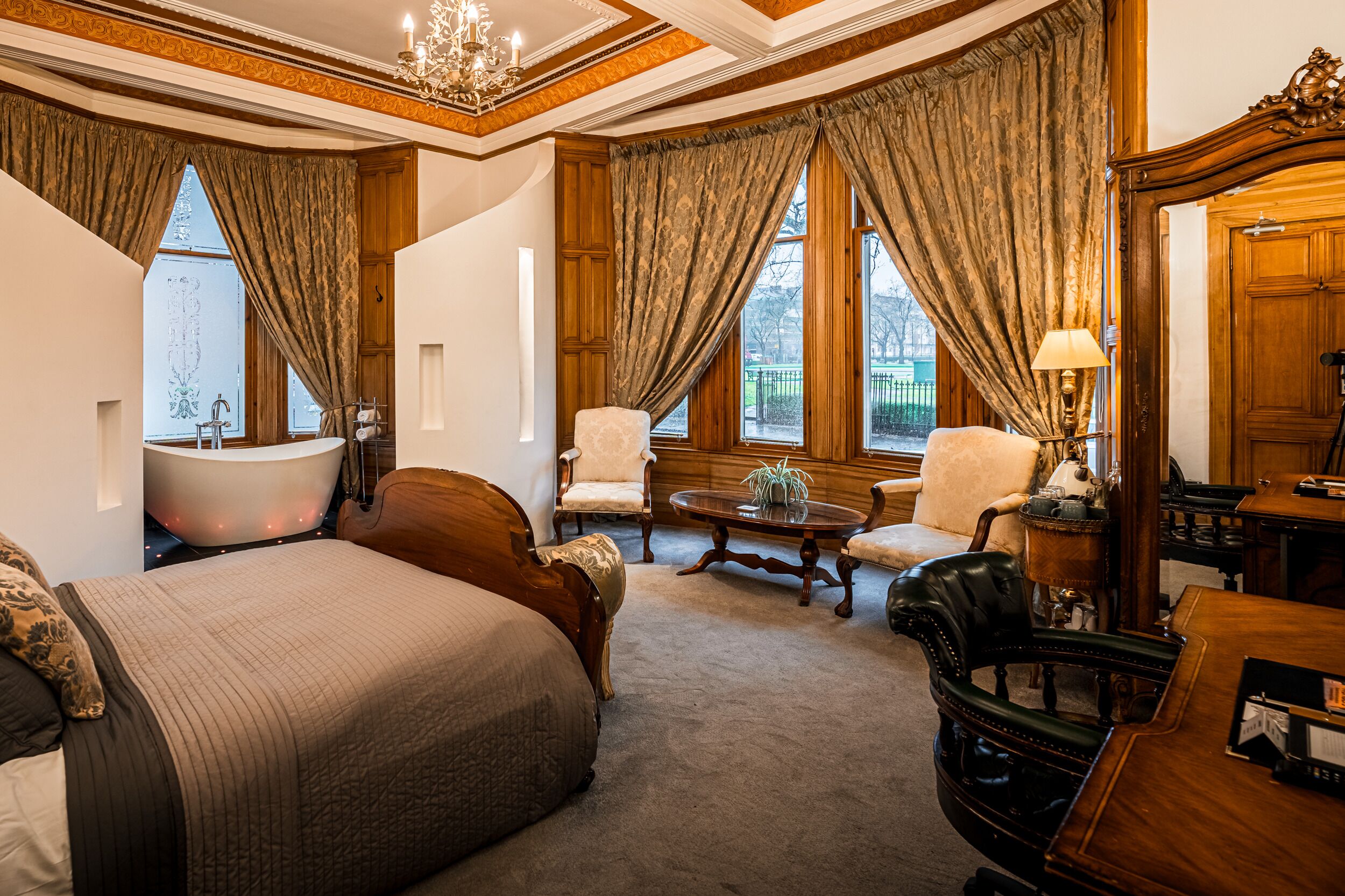 A spacious bedroom with a large bed, a sitting area with a table and chairs, a bathtub, and a large mirror.