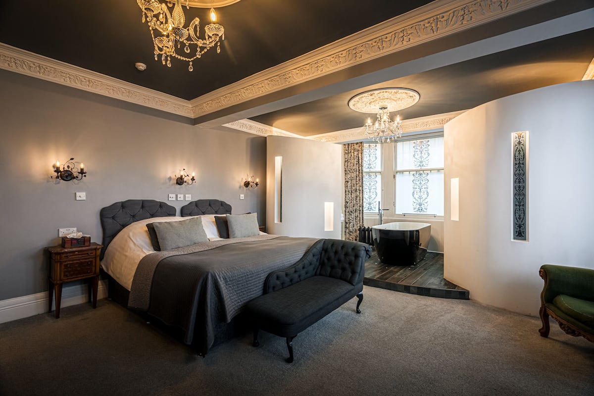 Une chambre à coucher avec un grand lit, un banc, une table de chevet, un lustre et une baignoire.