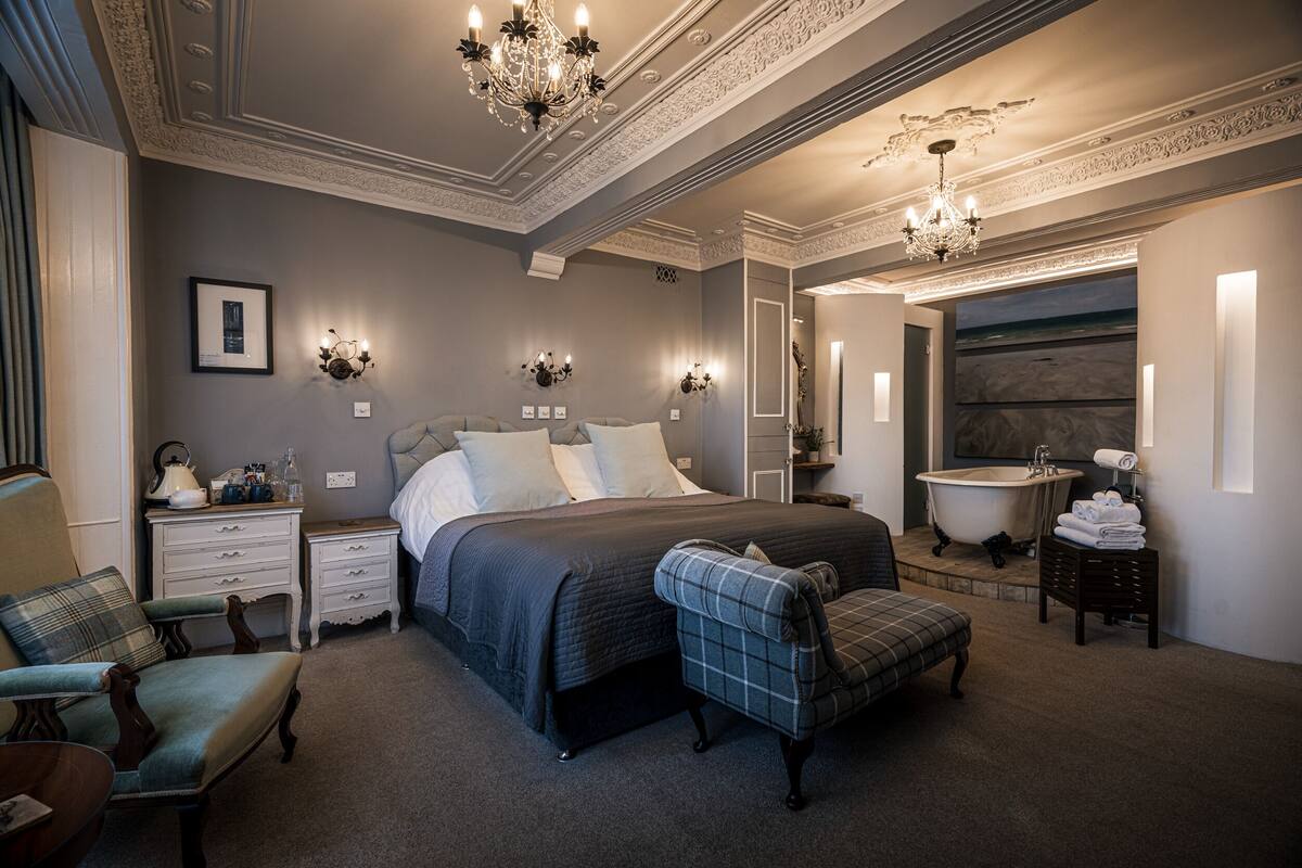 Une chambre à coucher avec un grand lit, des tables de chevet, une chaise, une coiffeuse avec un miroir et une baignoire.