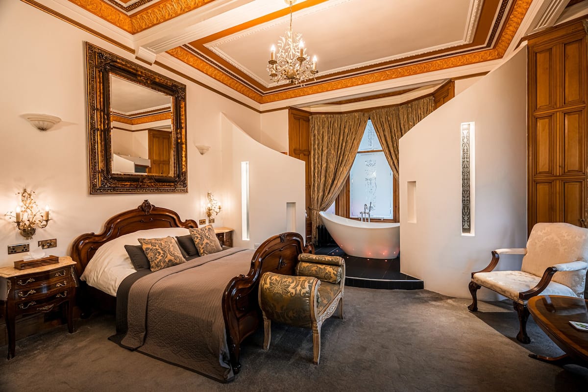 Une chambre spacieuse avec un grand lit, un coin salon avec un fauteuil, une coiffeuse et une baignoire.