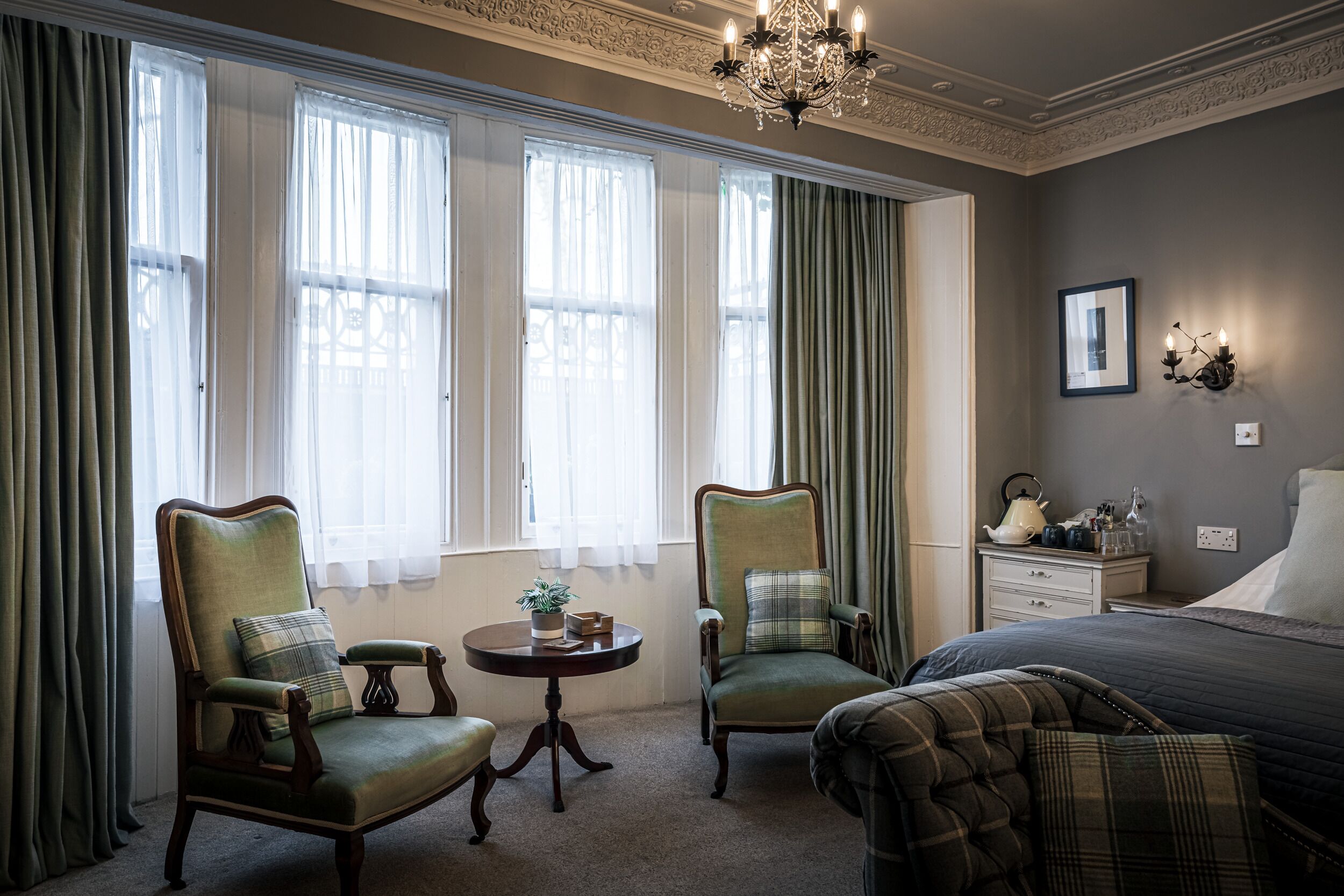 A room with two armchairs, a round table, a bed, and a nightstand with a lamp.