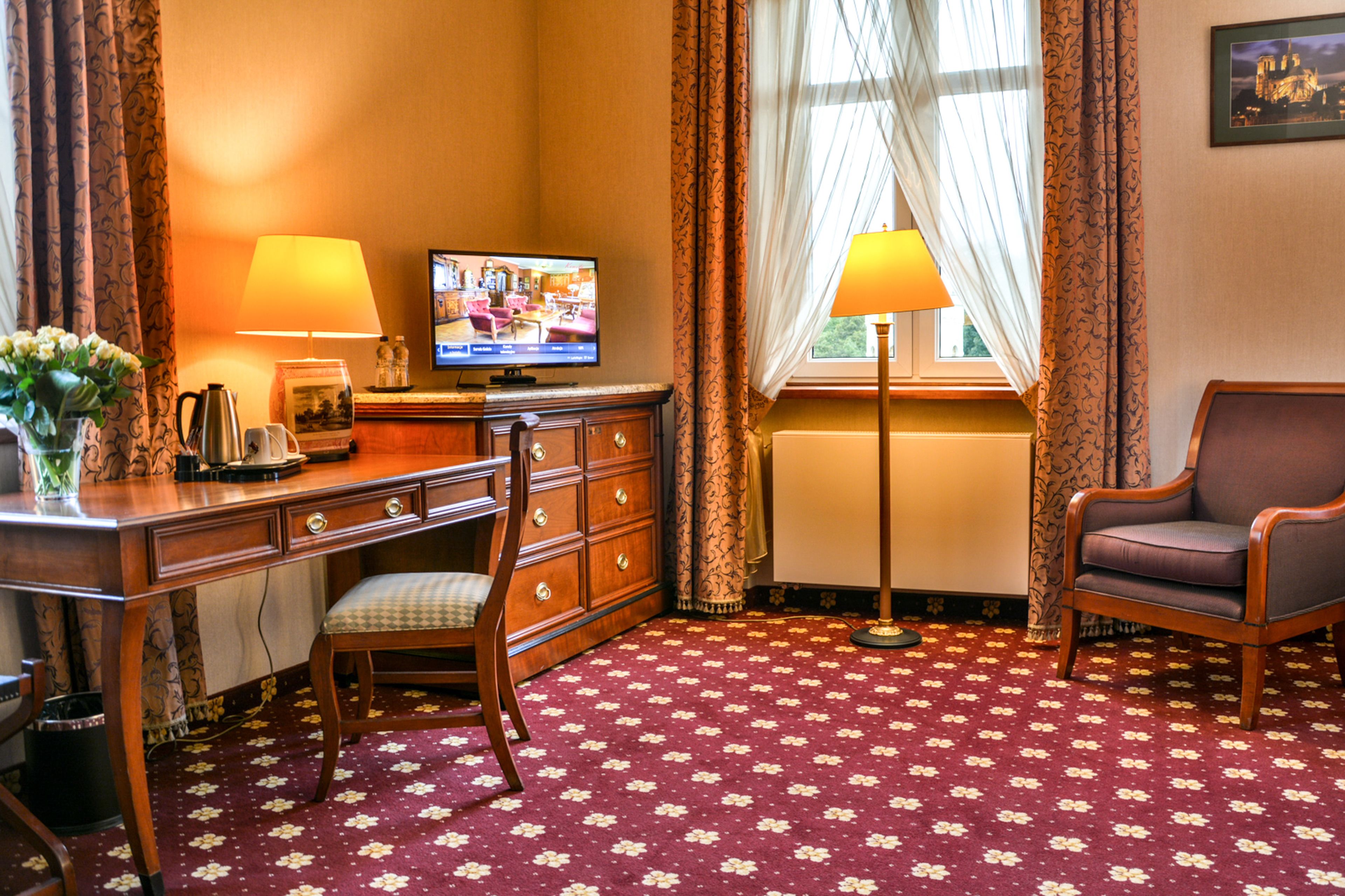 A hotel room with a wooden desk, a television, a chair, and a patterned carpet.