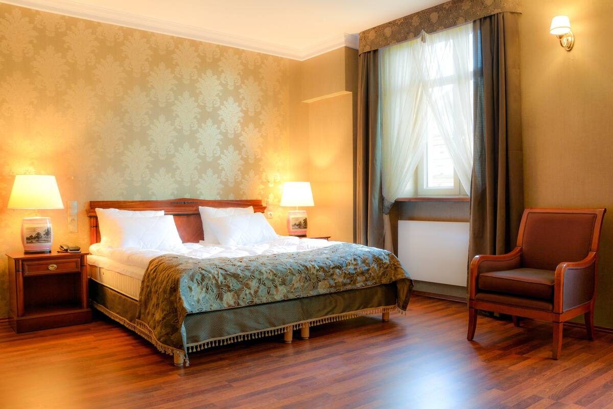 A hotel room with a large bed, two bedside tables with lamps, a chair, and a window with curtains.