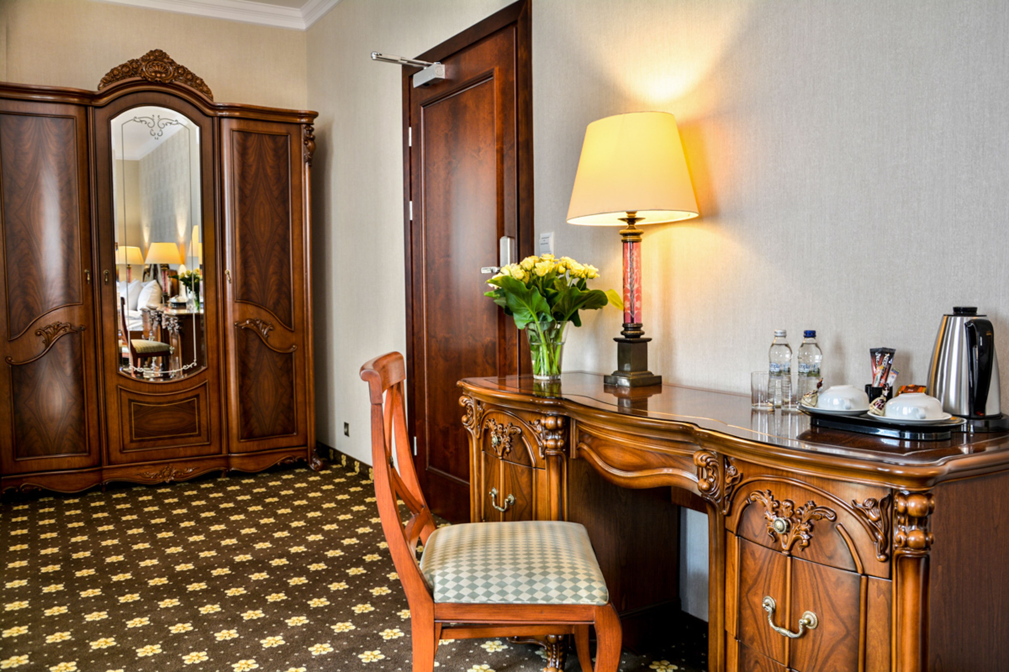 A hotel room with a wooden wardrobe, a desk with a lamp, a chair, and a mirror.