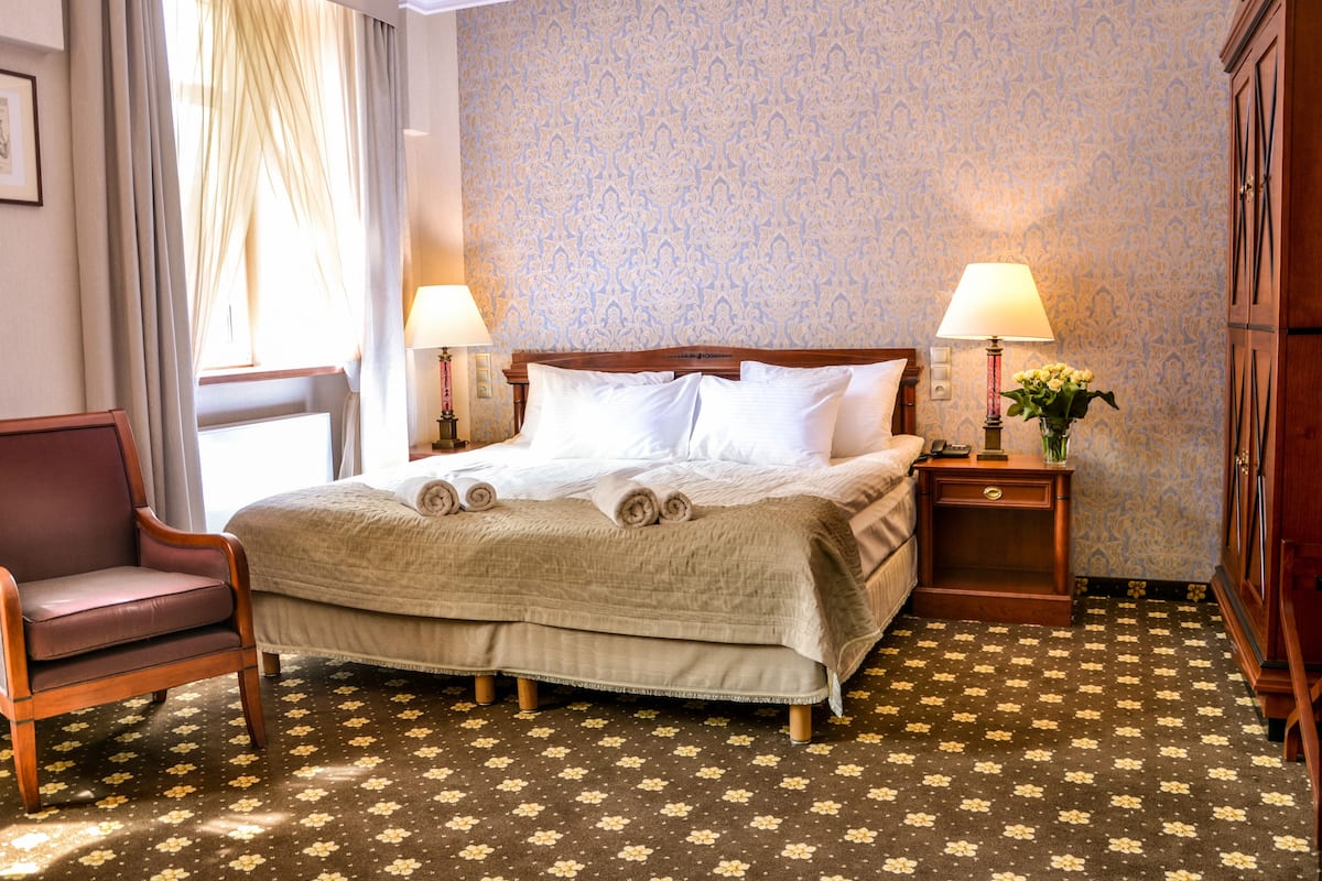 A neatly made bed with a wooden headboard, two bedside lamps, a floral-patterned carpet, and a wooden chair.