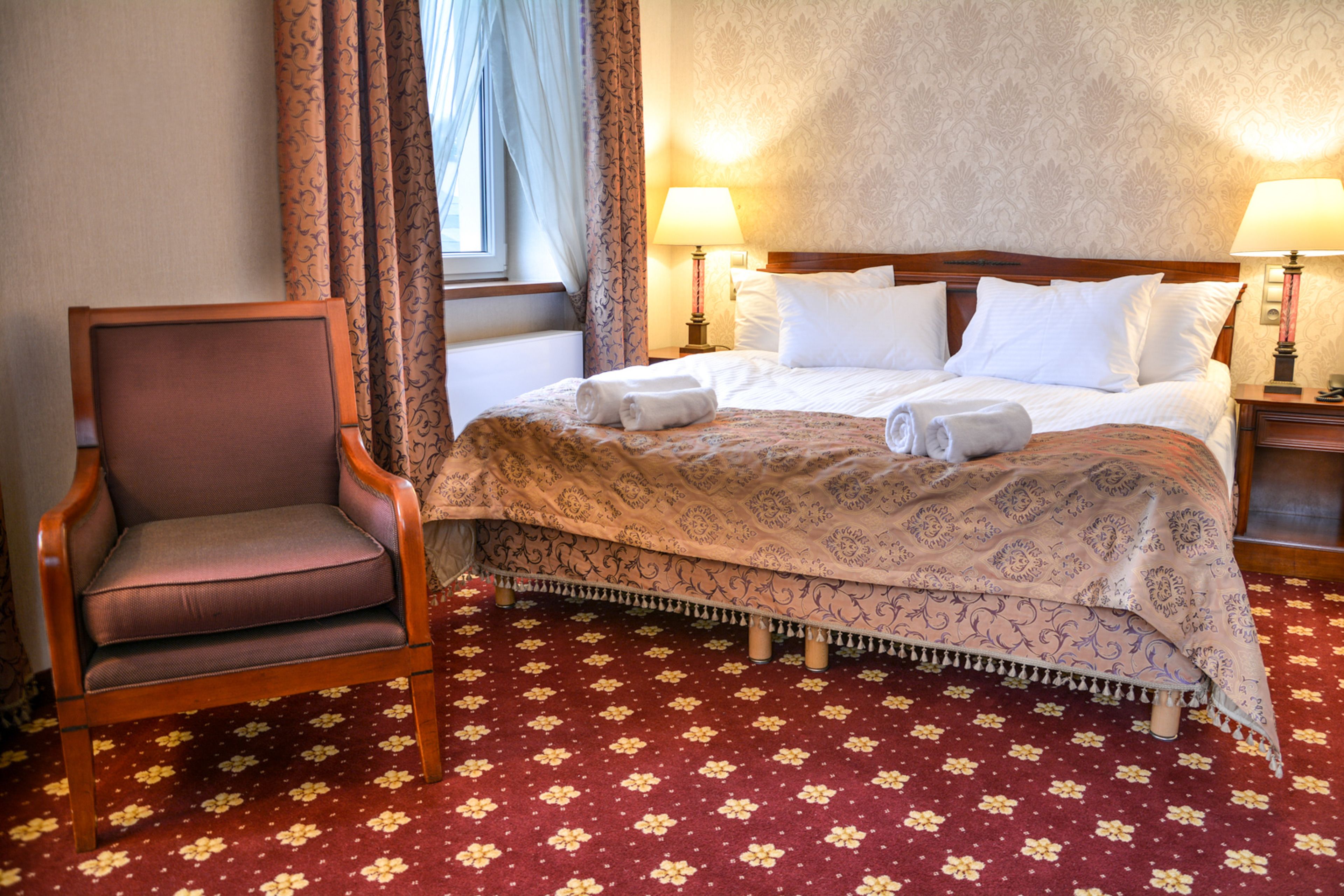 Une chambre d’hôtel avec un grand lit, deux lampes de chevet, une chaise en bois et une moquette à motifs floraux.