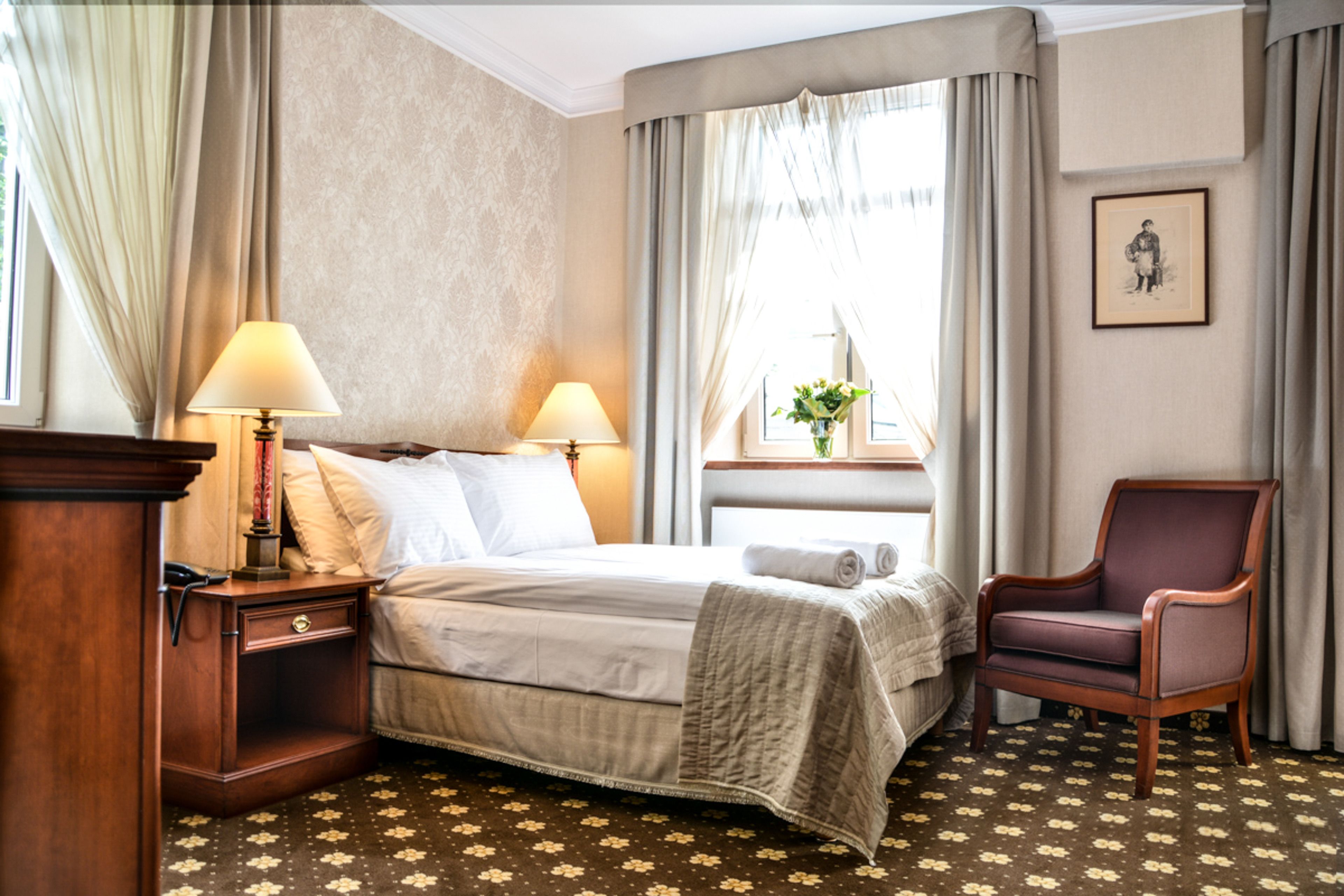 Et hotelværelse med en seng, to sengeborde, en lampe på hver natbord, en stol, et vindue med gardiner og et billede i ramme på væggen.