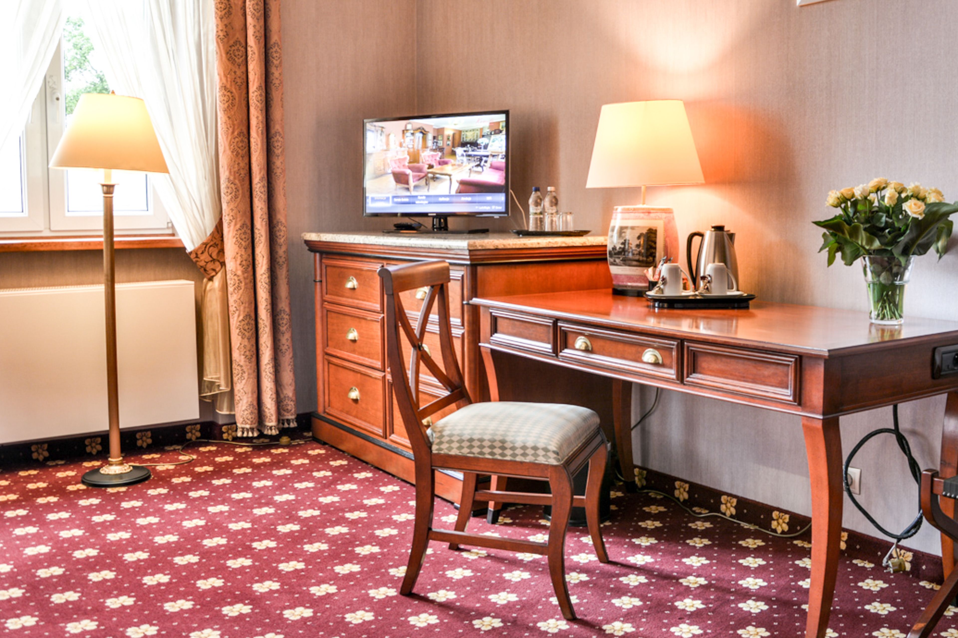 Habitación de hotel con escritorio de madera, una silla, televisor, dos lámparas, un jarrón con flores y alfombra estampada.