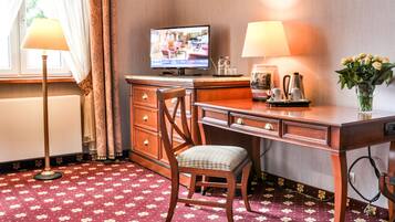 Habitación de hotel con escritorio de madera, una silla, televisor, dos lámparas, un jarrón con flores y alfombra estampada.