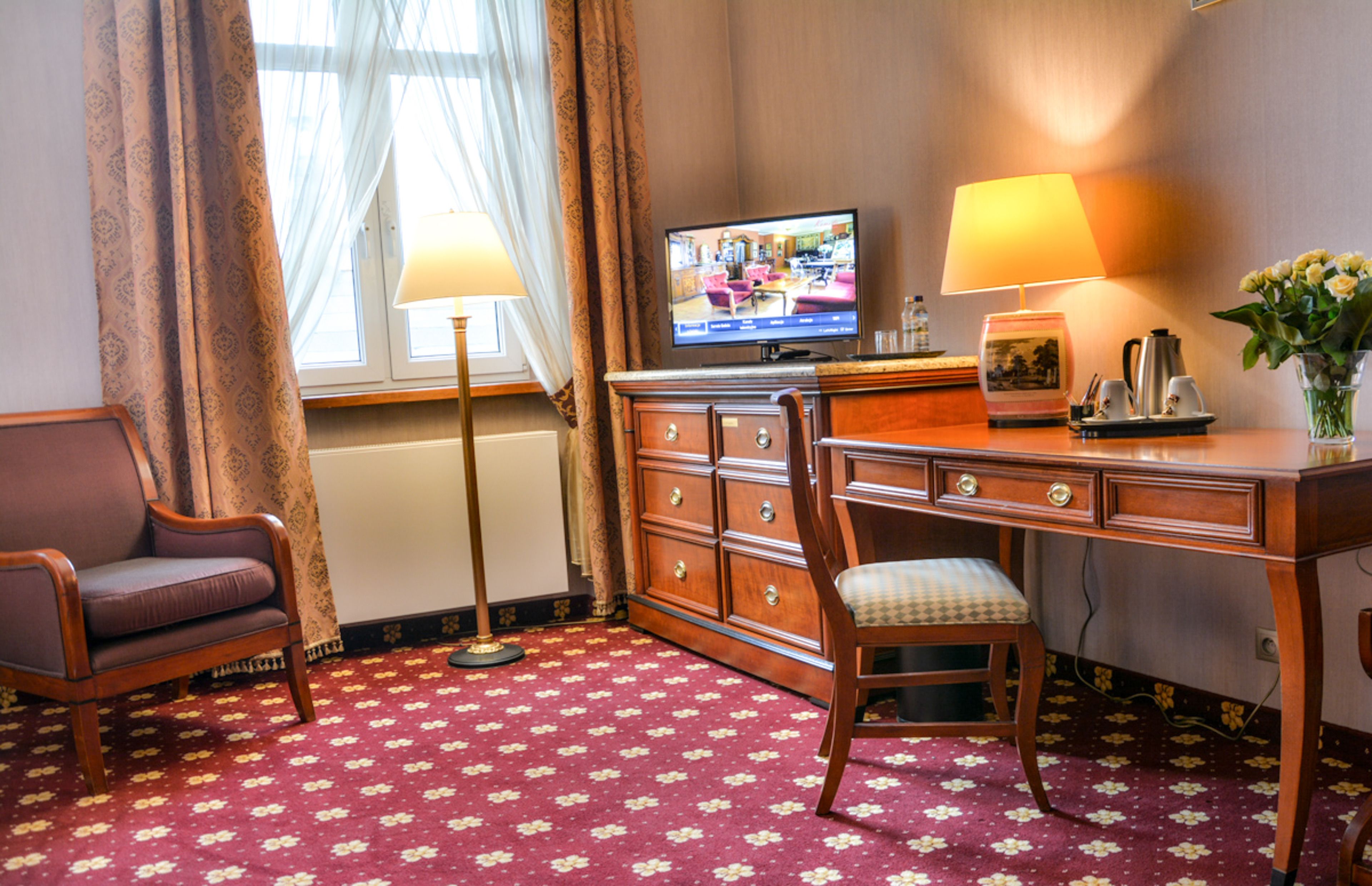 Ein Hotelzimmer mit einem Holztisch, einem Fernseher, einem Sessel, einer Lampe und einer Vase mit Blumen.