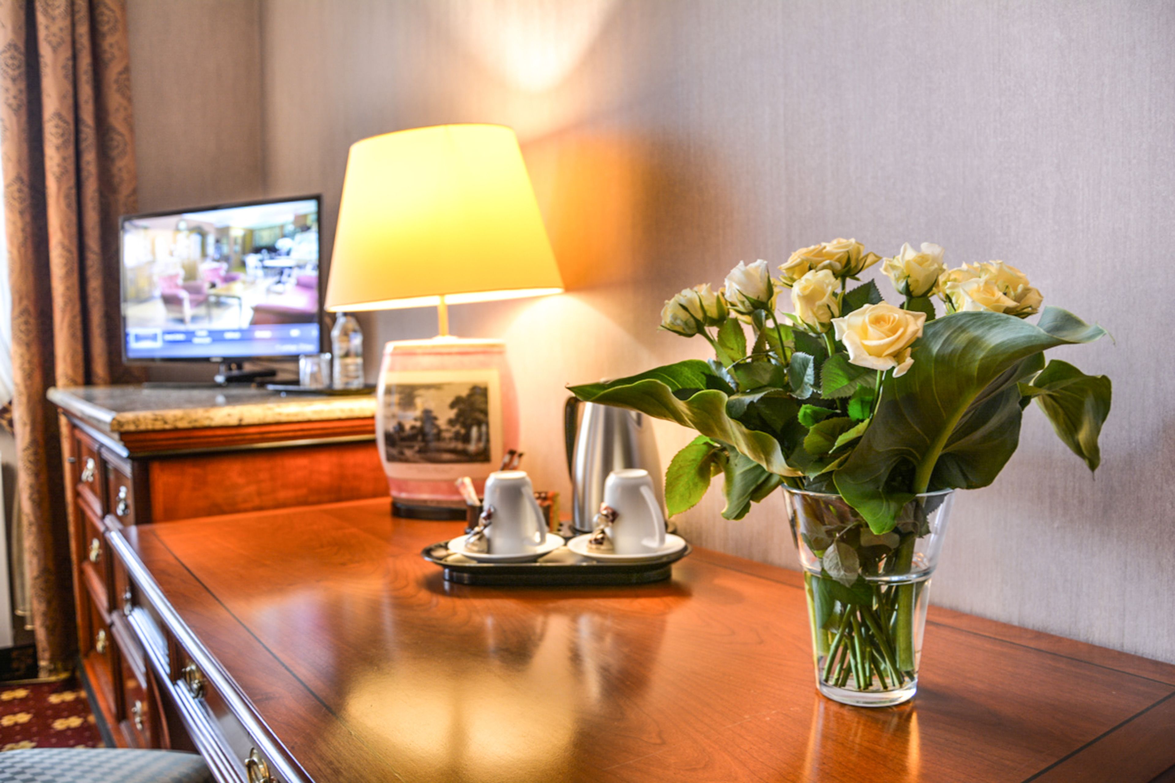 Ett träbord med en vas med blommor, en lampa och en tv-apparat i bakgrunden.