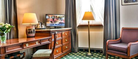 Habitación de hotel con un escritorio de madera, una silla, un televisor, una lámpara y una ventana con cortinas.