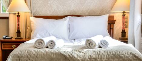 Una cama bien tendida con sábanas blancas y toallas enrolladas, un cabecero de madera y dos lámparas de noche con pantallas.