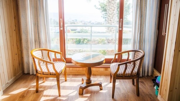 A room with two wicker chairs and a round glass table, large windows with curtains, and a view of greenery and a building outside. - Living Stone Pension (Seogwipo)