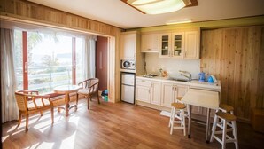 A compact kitchen with wooden cabinets, a refrigerator, and a microwave. There is a dining area with a round table and chairs. - Living Stone Pension (Seogwipo)