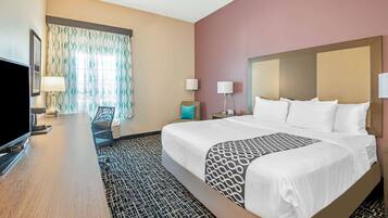 A hotel room with a large bed, a desk with a flat-screen TV, a chair, and a window with patterned curtains.