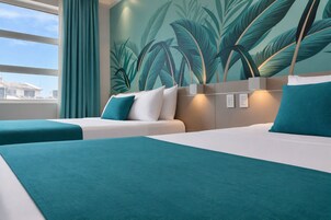 Una habitación de hotel con dos camas, ropa de cama color turquesa, un ventanal con cortinas y un mural de hojas tropicales en la pared.