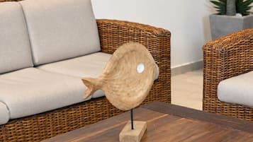 Um sofá de vime com almofada, uma escultura de peixe em madeira sobre uma mesa e uma planta em vaso ao fundo.