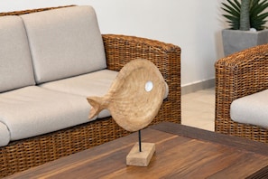 Un sofá de mimbre con un cojín, una escultura de pez de madera sobre una mesa y una planta en maceta al fondo.