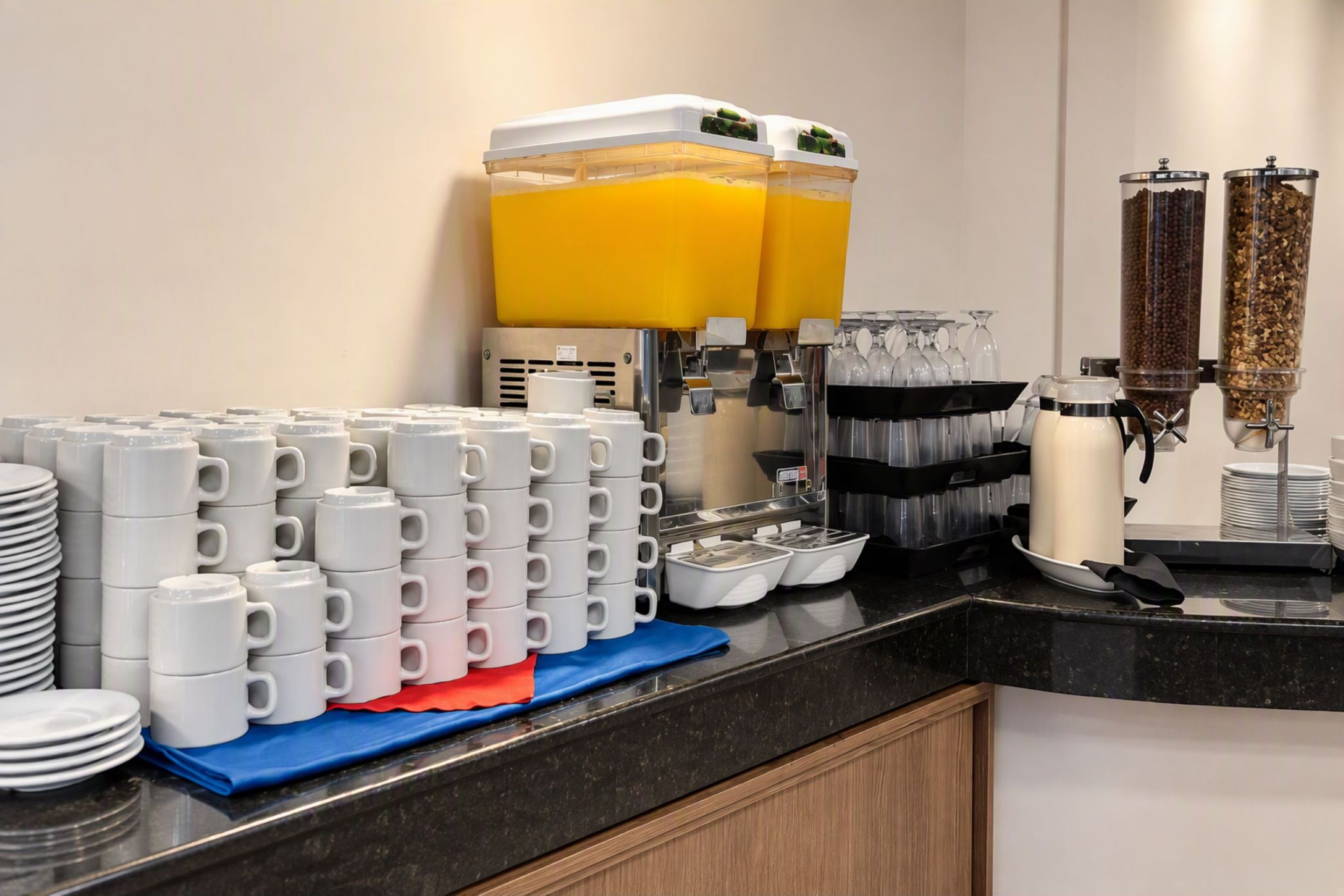 A breakfast buffet setup with stacked white mugs, juice dispensers, and a coffee machine.