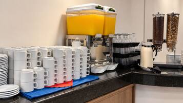 A breakfast buffet setup with stacked white mugs, juice dispensers, and a coffee machine.
