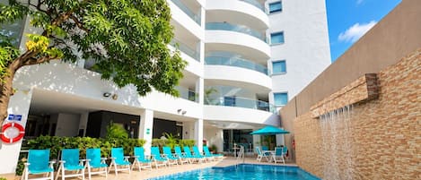 Una piscina con sillones de descanso, un edificio con varios balcones y un árbol.