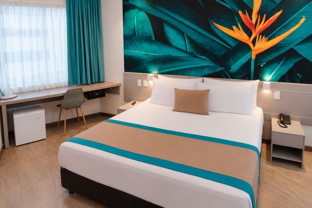 Habitación de hotel con cama, escritorio, silla, refrigerador pequeño y mesita de noche con teléfono.