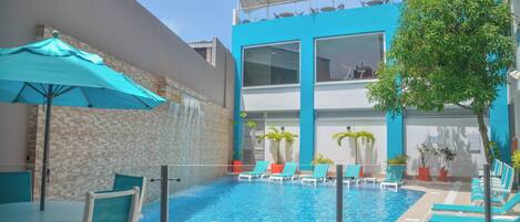 A swimming pool area with lounge chairs and umbrellas, a stone wall, and a modern building with large windows.