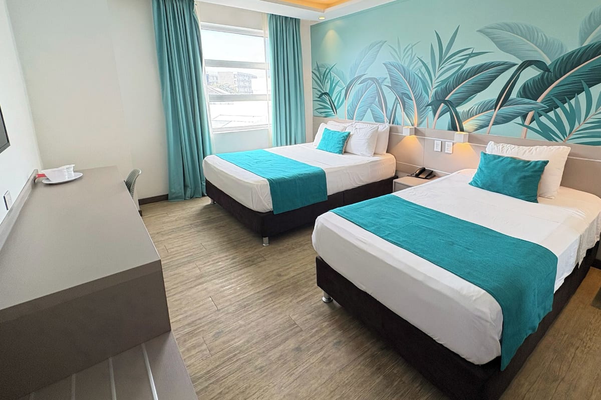 A hotel room with two beds, a desk, a TV, and a mural of tropical leaves.
