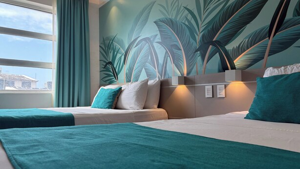 A hotel room with two beds, teal bedding, a large window with curtains, and a tropical leaf mural on the wall. - Hotel Portobelo Plaza las Ámericas (San Andrés)
