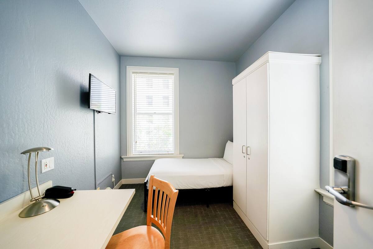 A small, well-lit room with a single bed, a desk, a chair, and a wardrobe.