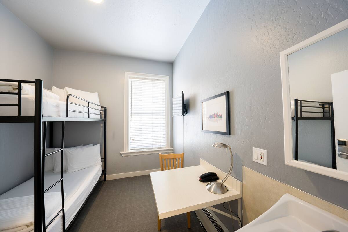 A compact hotel room with bunk beds, a desk, a chair, a desk lamp, a mirror, and a window with blinds.