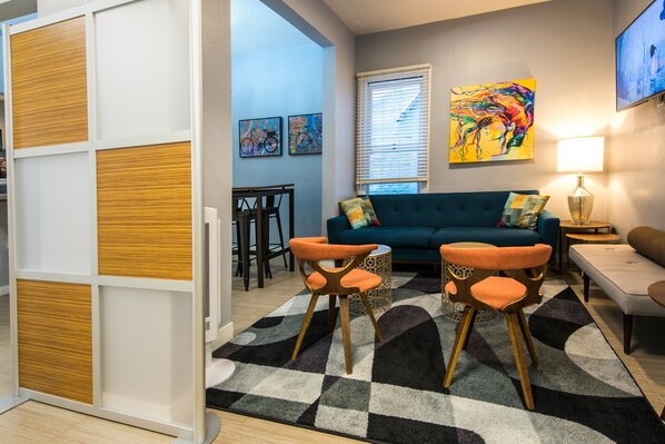 A modern living room with a teal sofa, orange chairs, a patterned rug, and artwork on the walls.