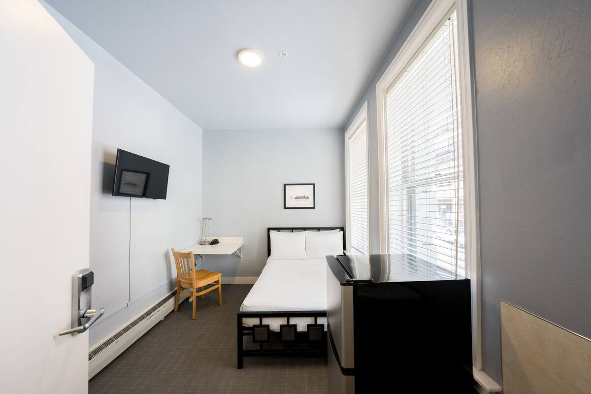 A compact hotel room with a single bed, a desk with a chair, a television, and a window with blinds.