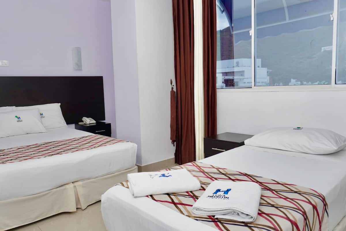 Habitación de hotel con dos camas, cada una con cubrecama blanco y manta a rayas. Se ve una ventana con cortinas marrones, una mesita de noche con teléfono y un interruptor de luz en la pared.