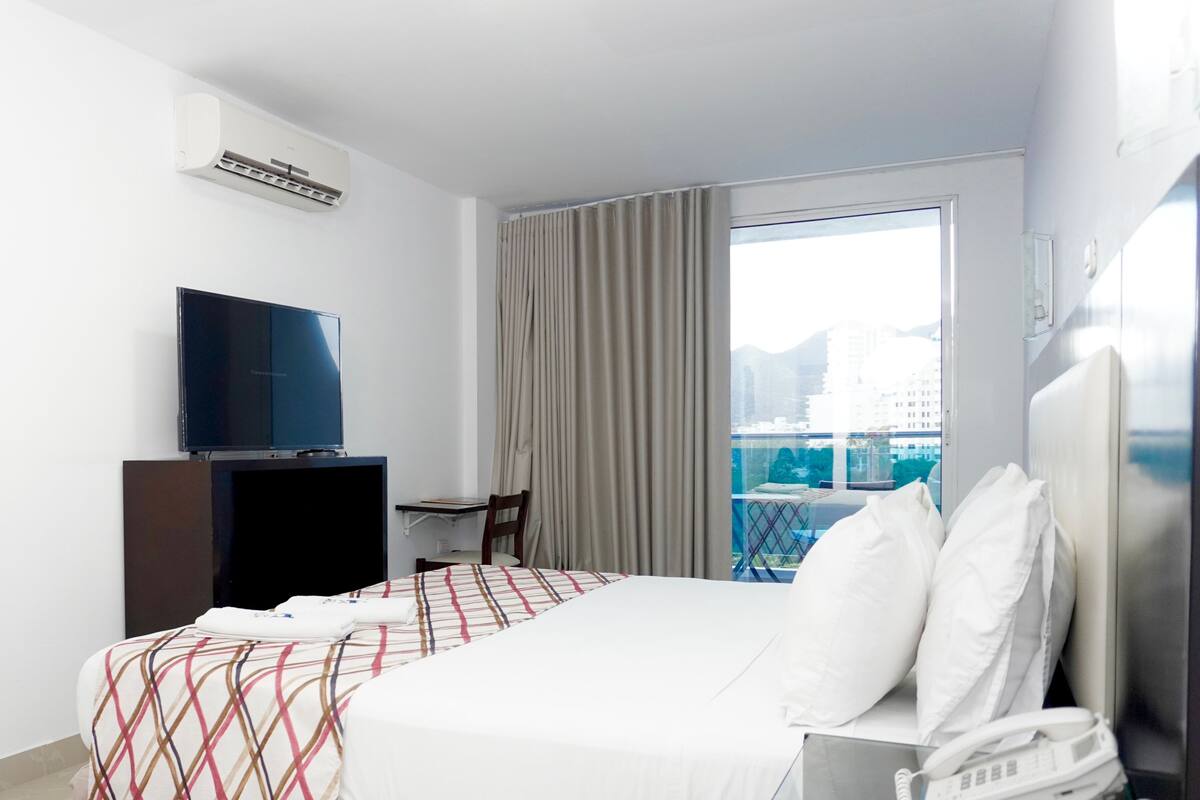 Habitación de hotel con cama, escritorio, televisión, teléfono y ventana con vista a edificios.