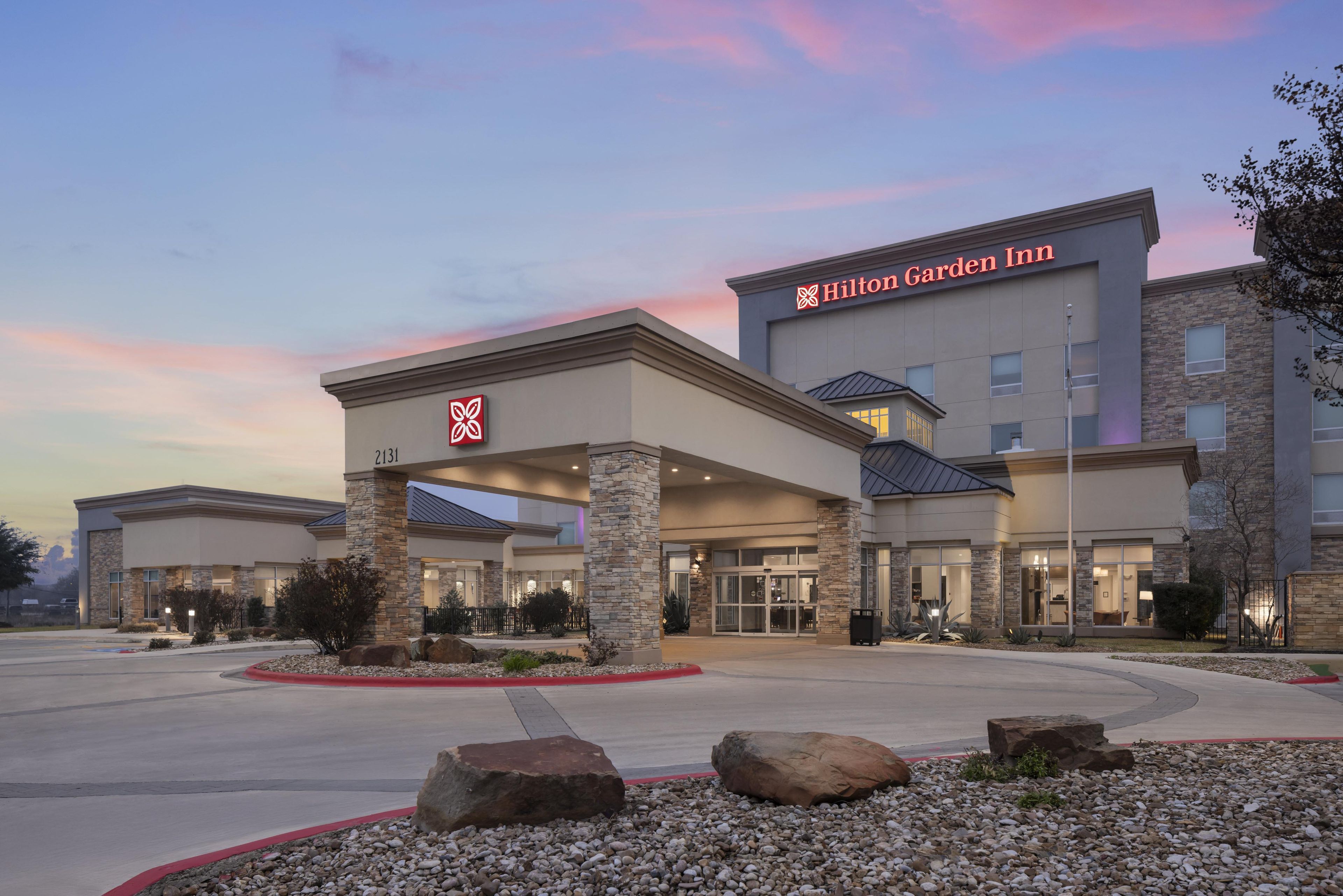 Un Hilton Garden Inn con un diseño arquitectónico moderno, destacando una entrada imponente, pilares de piedra y un área exterior bien cuidada con rocas y jardines.