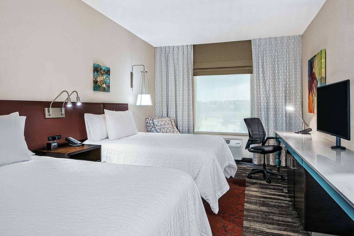 A hotel room with two beds, a desk with a computer, a chair, a TV, a window with curtains, and artwork on the walls.