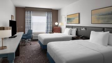 Habitación de hotel con dos camas, un escritorio con computadora, una silla, una ventana con cortinas, un cuadro en la pared y una mesita de noche con lámpara.