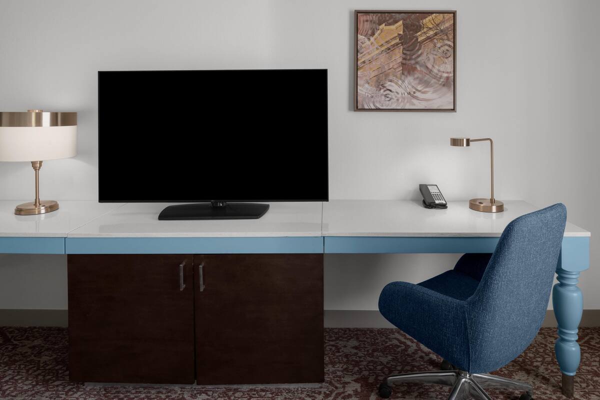 A hotel room with a large flat-screen TV, a desk with a lamp, a chair, and a cabinet.