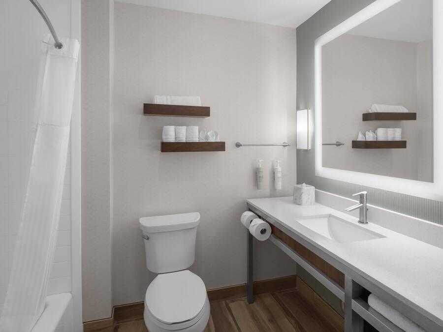 A modern bathroom with a toilet, sink, and shower. There are wooden shelves with towels and toiletries.