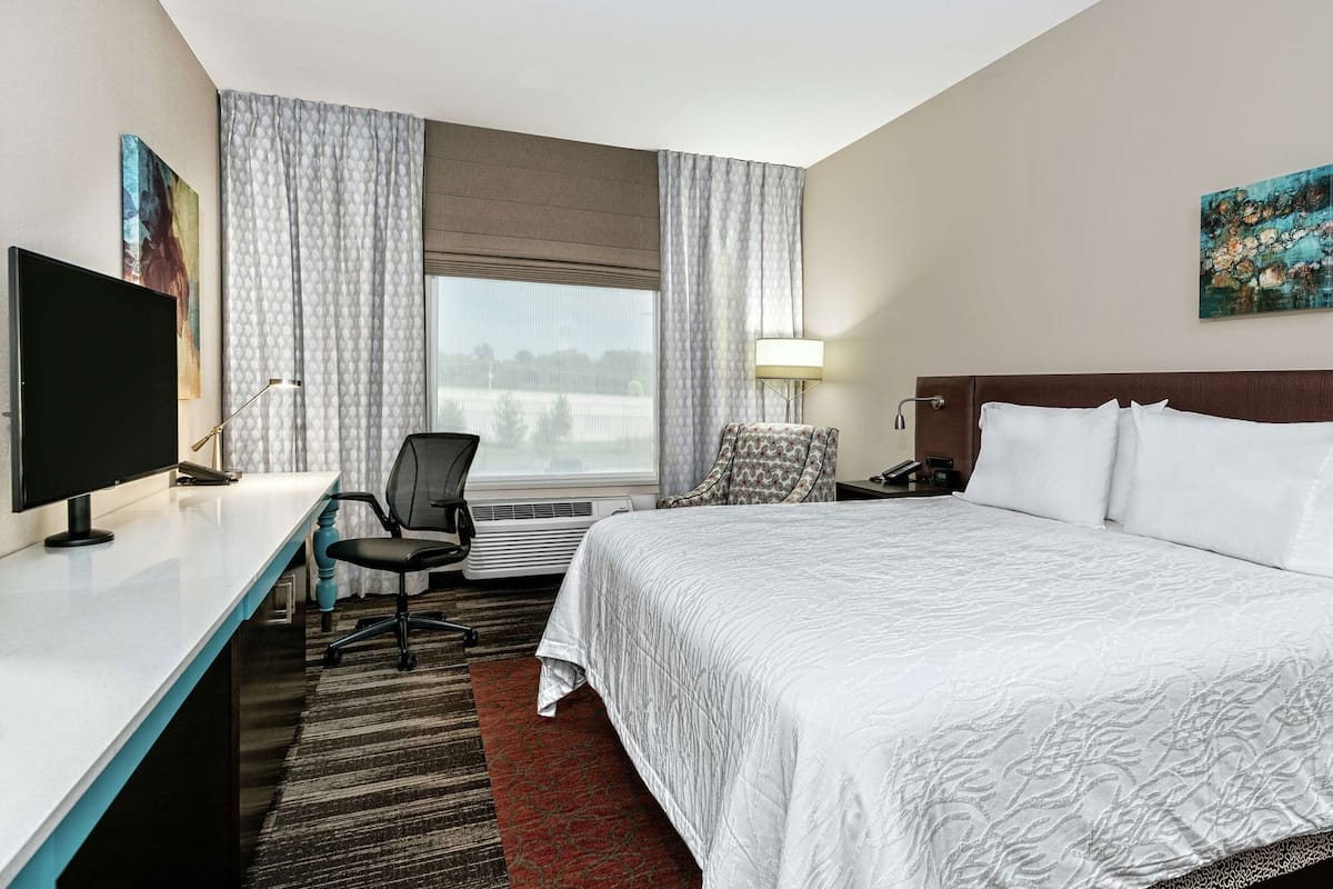 A hotel room with a bed, desk, chair, TV, and artwork.